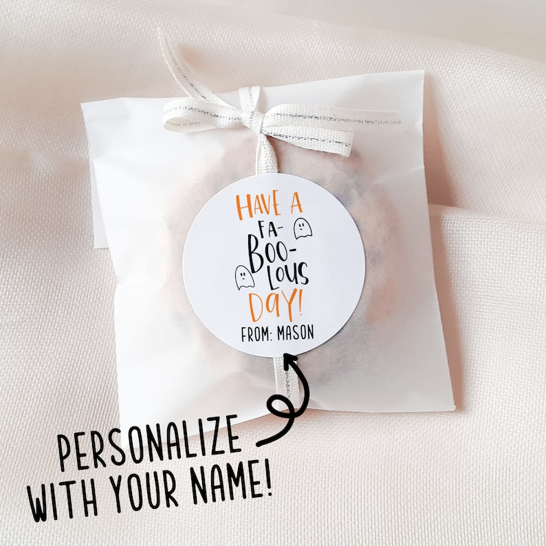 Op de afbeelding: Een witte ronde sticker met oranje tekst die "Have a Fa-Boo-Lous Day!" luidt, met twee geestillustraties. De tekst "From: Mason" staat onder de hoofdtekst. De sticker is bevestigd aan een witte papieren zak met een zilveren lint.