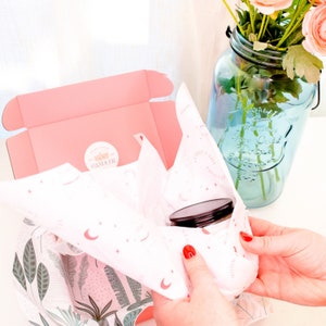 May include: A pink box with a white and pink patterned tissue paper inside. The tissue paper is partially pulled back to reveal a black candle jar with a black lid. The box has a sticker that says "CANDLER" on it.