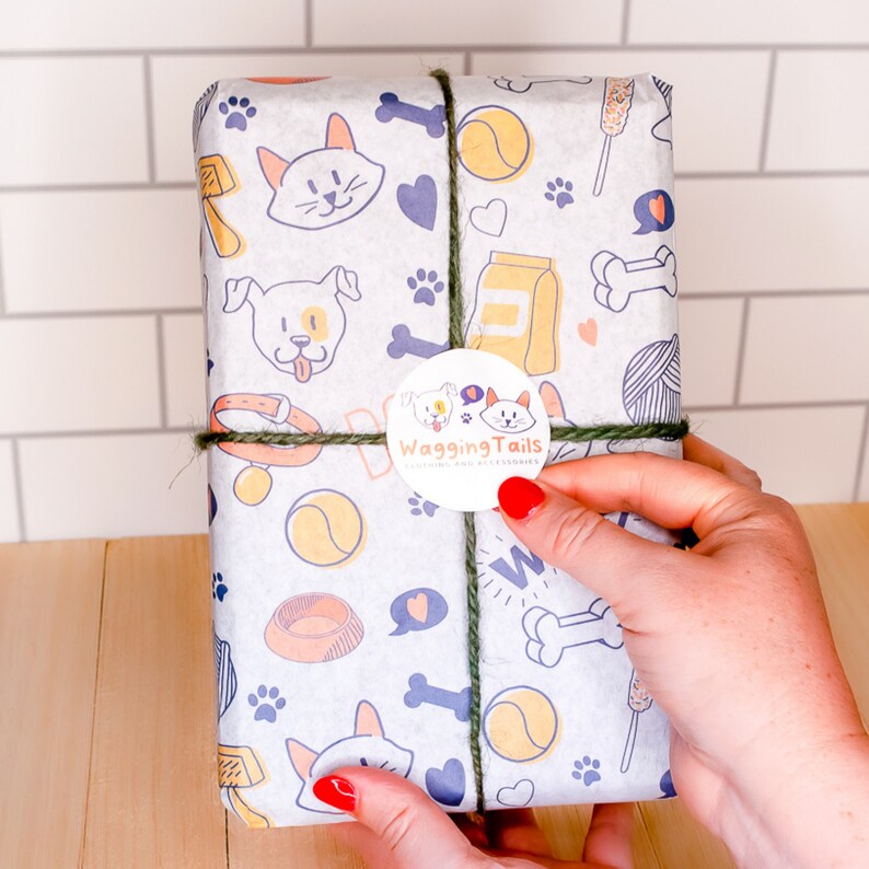 K&ouml;nnte beinhalten: Geschenk verpackt in wei&szlig;em Papier mit Hunde- und Katzenillustrationen, Pfotenabdr&uuml;cken und Icons zum Thema Haustiere. Das Paket ist mit gr&uuml;nem Garn verschn&uuml;rt und hat einen runden Aufkleber mit der Aufschrift "Wagging Tails".