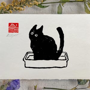 May include: A black cat illustration sitting in a litter box. The artwork is printed on white paper and features a red stamp with mountain and sun imagery. The cat has large, round eyes and a curved tail. The artist's signature is visible.