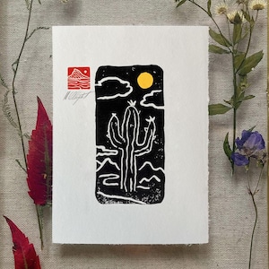 May include: A black and white linocut print of a cactus under a night sky with a yellow sun and clouds. The print has a red stamp and the artist's signature. The artwork is displayed on a white paper.