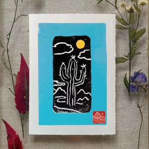 May include: A linocut print featuring a black cactus under a yellow sun and white clouds against a light blue background. The print is on white paper and includes a red mountain icon. Dried flowers and leaves surround the print.