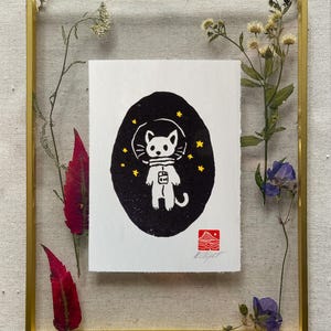 Puede incluir: Obra de arte enmarcada que muestra un gato astronauta blanco en un óvalo negro sobre fondo blanco. Estrellas doradas y un sello rojo acentúan la obra. El marco dorado contiene flores y hojas secas. El nombre del artista es visible.