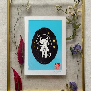May include: Framed art print featuring a white cat astronaut in a black oval against a light blue background, with yellow stars. The print is in a gold frame with dried flowers and leaves. The artist's name and the word "Art" are printed at the bottom.