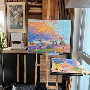 May include: A wooden easel with a colorful landscape painting on canvas. The painting depicts a mountain range with a bright orange and yellow sky. A black cat sits on a gray rug in the foreground.