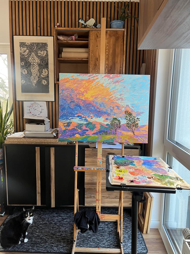 May include: A wooden easel with a colorful landscape painting on canvas. The painting depicts a mountain range with a bright orange and yellow sky. A black cat sits on a gray rug in the foreground.