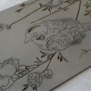 May include: A gray wooden plaque with a black line drawing of a bird perched on a branch with flowers. The bird is facing left and has a detailed design. The flowers are stylized and have a simple design.