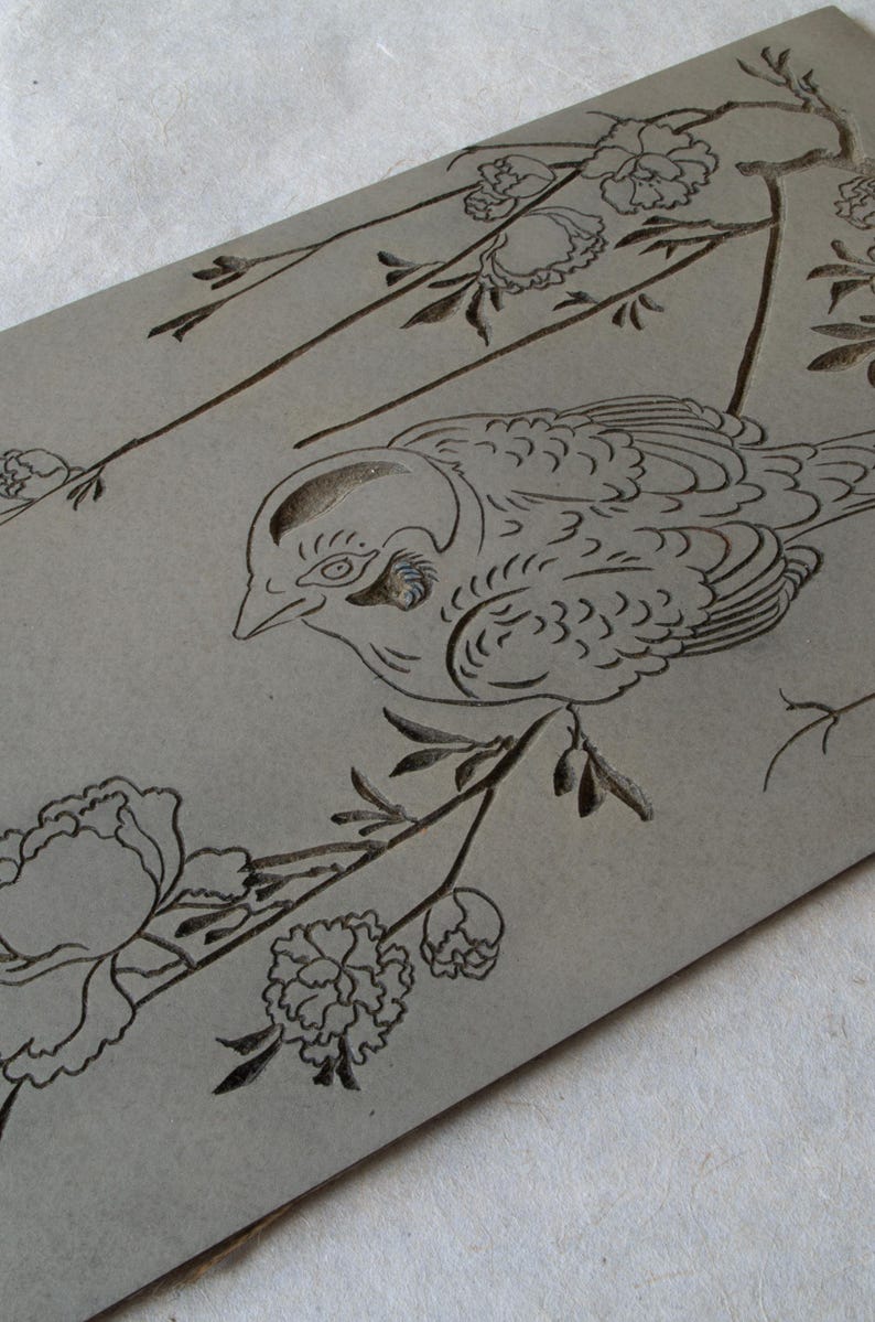 May include: A gray wooden plaque with a black line drawing of a bird perched on a branch with flowers. The bird is facing left and has a detailed design. The flowers are stylized and have a simple design.