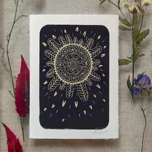 May include: A black and white art print featuring a sun design with intricate patterns. The sun is centered on a rectangular card with rounded corners. The print is surrounded by dried flowers and leaves.