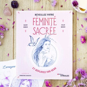 Peut inclure: Couverture de livre avec un fond rose et le titre "Réveillez Votre Féminité Sacrée" en grandes lettres roses et en gras. La couverture présente une illustration d'une femme aux longs cheveux flottants et d'un papillon. Le texte "Et Déployez Vos Sens" se trouve sous l'illustration. Le livre est de Daisy Bodin et Julie Bodin, avec des illustrations de Christine Chauvey. Le livre est publié par Editions Eyrolles.