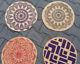 Op de afbeelding: Vier ronde jute placemats met decoratieve ontwerpen. Eén heeft een paars bloemenpatroon, een andere een zwart bloemendesign, een derde een rood bloemenpatroon en de laatste een paars geometrisch patroon. De placemats zijn gerangschikt op een grijze ondergrond.