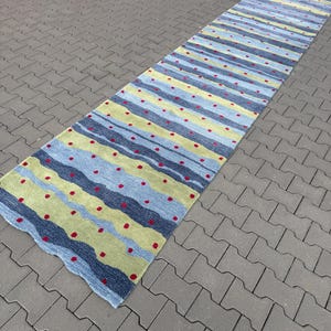 Op de afbeelding: Een lange, rechthoekige loper met een golvend, horizontaal streeppatroon in tinten lichtblauw, donkerblauw en limoengroen. Het oppervlak is versierd met kleine, gelijkmatig verdeelde rode cirkelvormige stippen over de gehele lengte van het kleed.