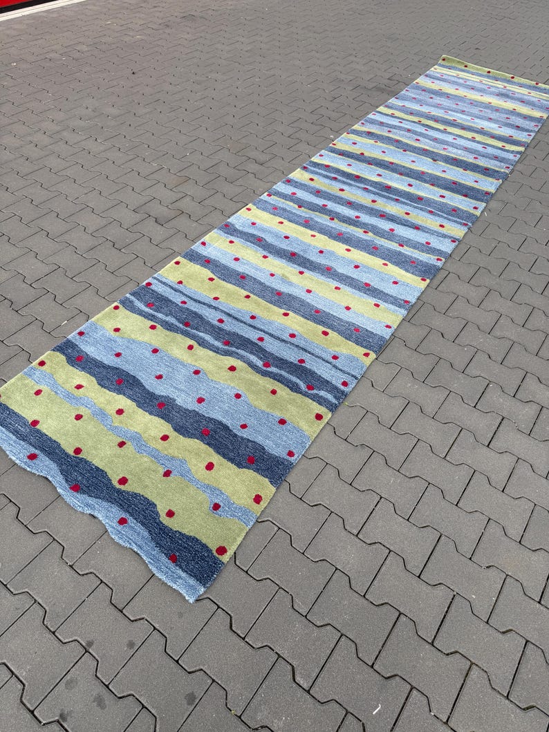 Op de afbeelding: Een lange, rechthoekige loper met een golvend, horizontaal streeppatroon in tinten lichtblauw, donkerblauw en limoengroen. Het oppervlak is versierd met kleine, gelijkmatig verdeelde rode cirkelvormige stippen over de gehele lengte van het kleed.