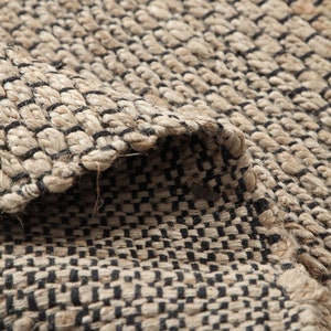 May include: Close-up of a woven rug with a black and beige chequered pattern. The rug is made of natural fibres and has a rough texture.