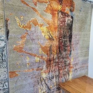 May include: Abstract rug with a gray background and orange, yellow, and blue abstract designs. The rug is hanging on a wall.