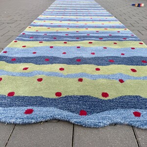 Op de afbeelding: Een lang rechthoekig loperkleed met horizontale golvende banen in tinten lichtblauw, donkerblauw en limoengroen. Het kleed is versierd met een herhalend patroon van kleine, effen rode cirkelvormige stippen verspreid over het gehele oppervlak.