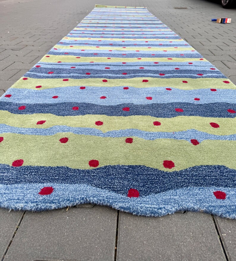 Op de afbeelding: Een lang rechthoekig loperkleed met horizontale golvende banen in tinten lichtblauw, donkerblauw en limoengroen. Het kleed is versierd met een herhalend patroon van kleine, effen rode cirkelvormige stippen verspreid over het gehele oppervlak.