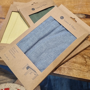 Könnte beinhalten: Drei Tischläufer in braunen Schachteln mit einem Fenster, das den Stoff zeigt. Die Tischläufer sind in Blau, Grün und Gelb. Die Schachteln tragen den Text "Linen & More" und ein Logo mit dem Buchstaben "M". Der blaue Tischläufer trägt den Text "Table runner, Tafelloper, Tischläufer, Chemin de table, 50 x 140 cm".