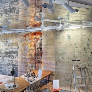 May include: Three large, abstract patterned rugs hang on a wall in a store. The rugs are in shades of beige, gray, and brown with hints of red and green. A wooden table sits in front of the rugs with a few decorative items on top.