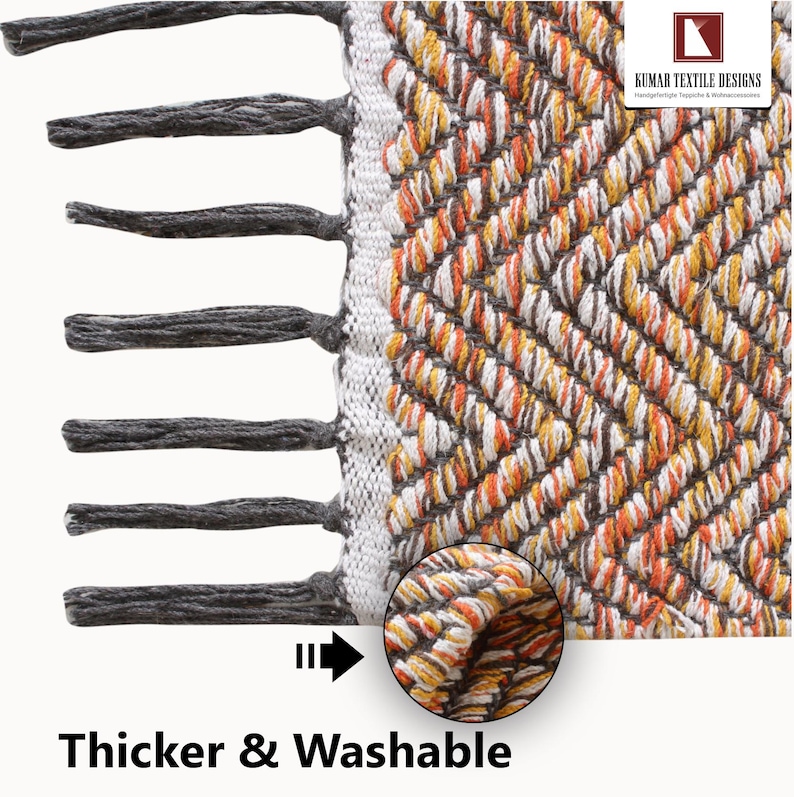 K&ouml;nnte beinhalten: Nahaufnahme eines gewebten Teppichs mit einem geometrischen Muster in Orange-, Wei&szlig;- und Braunt&ouml;nen. Der Teppich hat einen Fransenrand aus grauen Quasten an einer Kante. Der Text "Thicker & Washable" steht unten.