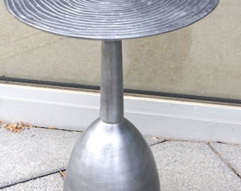 Op de afbeelding: Een zilveren metalen bijzettafel met een rond blad en een uniek, getextureerd, spiraalvormig ontwerp. De tafel heeft een hoge, slanke basis in de vorm van een vaas.