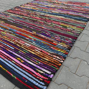 Peut inclure: Un tapis rectangulaire multicolore avec un motif rayé. Le tapis présente une variété de couleurs, notamment le rouge, le bleu, le violet et l'orange. Les bords sont finis avec une bordure noire. Le tapis est en tissu.