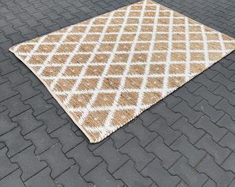 Op de afbeelding: Rechthoekig tapijt met een ruitpatroon. Het tapijt heeft een natuurlijke bruine kleur met witte ruitvormen. Het tapijt is gemaakt van geweven materiaal en ligt op een grijs bakstenen oppervlak.