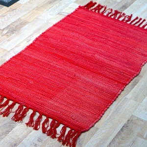 May include: A rectangular red woven rug with fringed edges. The rug is made of a tightly woven material and has a solid red colour. The rug is placed on a light-coloured wooden floor.