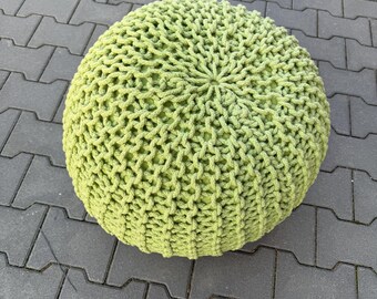 Op de afbeelding: Een ronde, limoengroene gebreide poef. De poef is gemaakt van dik garen, met een getextureerd, open weefpatroon. De poef staat op een grijs, geplaveid oppervlak. De poef is een decoratief en functioneel meubelstuk.