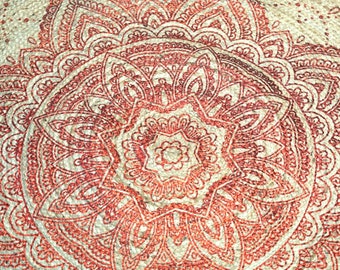 May include: A close-up of a cream-colored fabric with a red mandala design. The mandala is intricate and detailed, with a variety of shapes and patterns.