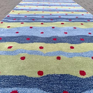 Op de afbeelding: Een lang, rechthoekig loperkleed met een herhalend patroon van horizontale golvende strepen in tinten lichtblauw, donkerblauw en limoengroen. Het oppervlak is over de gehele lengte versierd met kleine, gelijkmatig verdeelde rode stippen.
