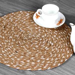 Op de afbeelding: Een ronde, bruin-witte geweven jute placemat met een witte rand. De placemat ligt op een grijs houten oppervlak. Een wit theekopje en schotel met bloemenmotieven staan op de placemat. Een witte theepot staat aan de rechterkant van de afbeelding.