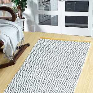 Peut inclure: Un tapis blanc et gris à motifs géométriques avec une bordure jaune. Le tapis est posé sur un sol en bois clair.