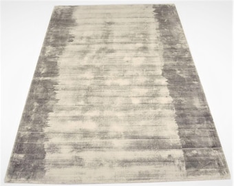 Op de afbeelding: Een rechthoekig vloerkleed met een distressed, abstract ontwerp. Het vloerkleed heeft een lichtbeige midden met donkerdere grijze randen en accenten, waardoor een textuurrijk, schilderachtig effect ontstaat. De afmetingen van het vloerkleed zijn niet zichtbaar op de afbeelding.