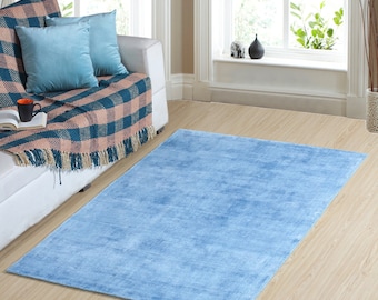 Op de afbeelding: Een rechthoekig, lichtblauw tapijt op een houten vloer. Het tapijt ligt voor een witte bank met turquoise kussens en een geruite plaid. Op de achtergrond is een raam met natuurlijk licht.