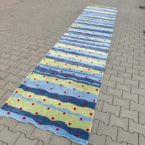 Op de afbeelding: Een lang rechthoekig loperkleed met horizontale golvende strepen in tinten lichtblauw, donkerblauw en limoengroen. Het oppervlak is versierd met een herhalend patroon van kleine, gelijkmatig verdeelde rode stippen over de gehele lengte.
