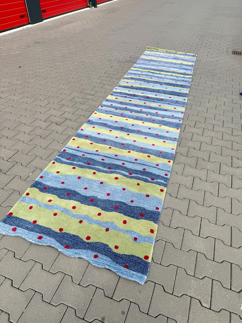 Op de afbeelding: Een lang rechthoekig loperkleed met horizontale golvende strepen in tinten lichtblauw, donkerblauw en limoengroen. Het oppervlak is versierd met een herhalend patroon van kleine, gelijkmatig verdeelde rode stippen over de gehele lengte.