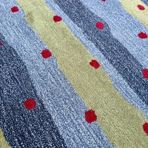 Op de afbeelding: Een getextureerd tapijt met diagonale strepen in afwisselende tinten lichtblauw, donkerblauw en saliegroen. Het oppervlak is versierd met een herhalend patroon van kleine, effen rode cirkelvormige stippen verspreid over het gehele oppervlak.