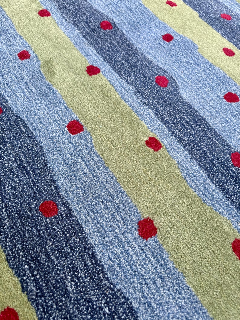 Op de afbeelding: Een getextureerd tapijt met diagonale strepen in afwisselende tinten lichtblauw, donkerblauw en saliegroen. Het oppervlak is versierd met een herhalend patroon van kleine, effen rode cirkelvormige stippen verspreid over het gehele oppervlak.