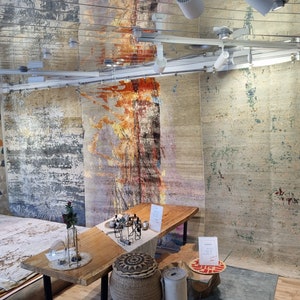 May include: A display of colorful, abstract patterned rugs in a showroom setting. The rugs are hanging on the wall and laid out on the floor. A wooden table with a vase of flowers sits in the center of the room.
