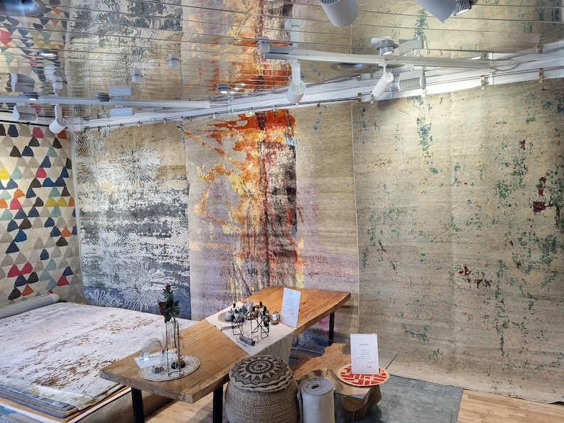 May include: A display of colorful, abstract patterned rugs in a showroom setting. The rugs are hanging on the wall and laid out on the floor. A wooden table with a vase of flowers sits in the center of the room.