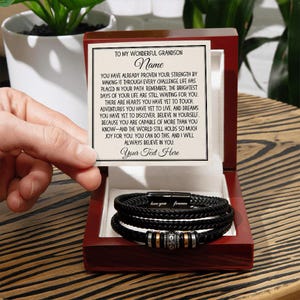 Puede incluir: Una pulsera de cuero trenzado negro con detalles plateados y dorados, presentada en una caja de madera roja. La caja contiene una tarjeta personalizada con el texto "TO MY WONDERFUL GRANDSON" y espacio para un nombre y texto personalizado. La pulsera tiene las palabras "love you forever".