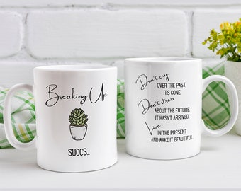 May include: Two white ceramic mugs with black text and a small green succulent illustration. The mug on the left says "Breaking Up SUCCS." The mug on the right says "Don't cry over the past. It's gone. Don't stress about the future. It hasn't arrived. Live in the present and make it beautiful."