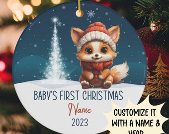 Può includere: Un addobbo natalizio rotondo, blu e bianco con una volpe di cartone animato che indossa un cappello rosso e una sciarpa. L'addobbo ha il testo "Baby's First Christmas" e uno spazio per personalizzare con un nome e un anno. L'anno 2023 è stampato sull'addobbo.