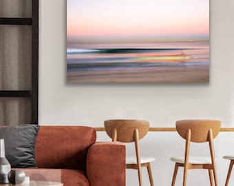 May include: A blurred photograph of a beach scene at sunset. The sky is a soft pink and purple, and the water is a deep blue. The sand is a light brown, and there is a faint outline of a person in the distance.
