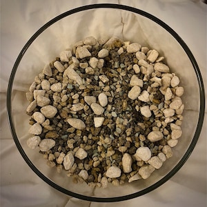 May include: A glass bowl filled with a variety of small, light brown and gray stones. The stones are arranged in a random pattern and appear to be natural.