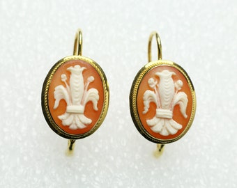 May include: A pair of gold-tone earrings featuring oval-shaped cameos. Each cameo has a light orange background with a white floral design. The earrings have a lever-back closure.