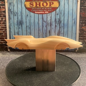May include: A wooden model of a classic car, unfinished, with a streamlined design. The car is mounted on a rectangular wooden base. The background features a sign that reads "DAD'S HOT ROD SHOP" in red and yellow.