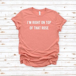 Puede incluir: Una camiseta de color coral con el texto "I'm right on top of that rose" impreso en blanco.