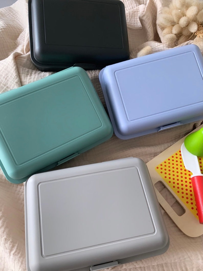 May include: Four plastic lunch boxes in different colors: black, light blue, light green, and gray. The boxes are all closed and sitting on a light brown fabric surface.